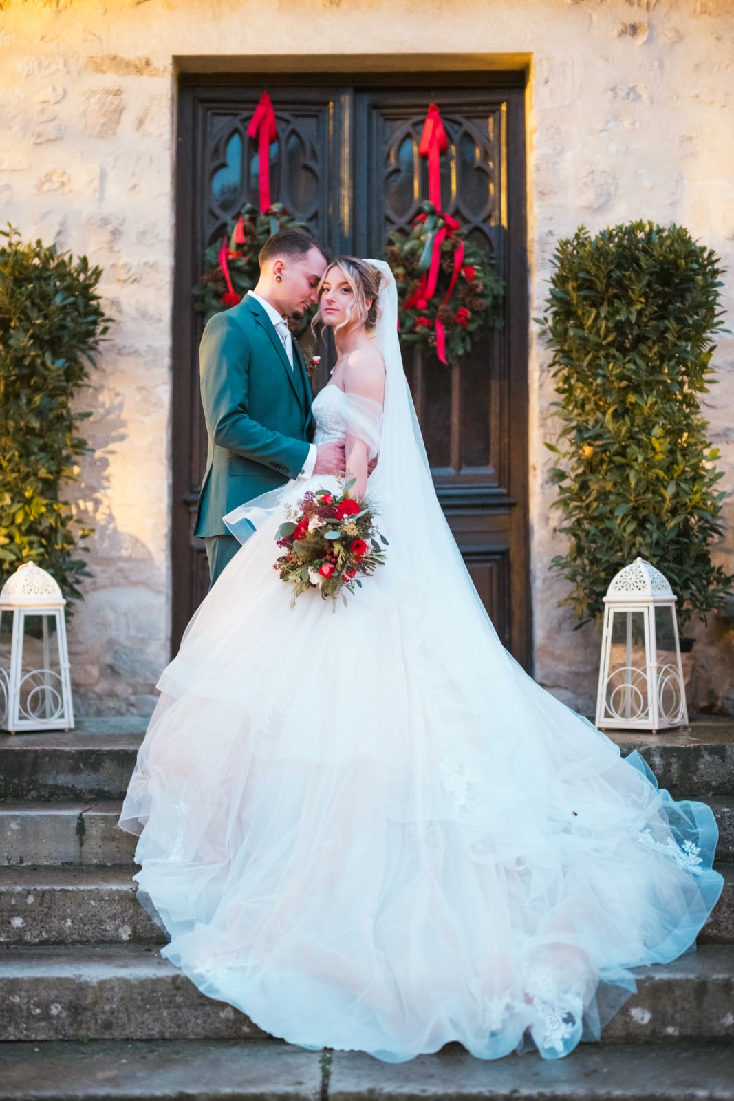 Couple devant la grande porte en bois sculptée avec couronnes de Noël aux rubans rouges, mariage au Château de Pontarmé, par Jean-Baptiste Argyre (Jaysphotography)