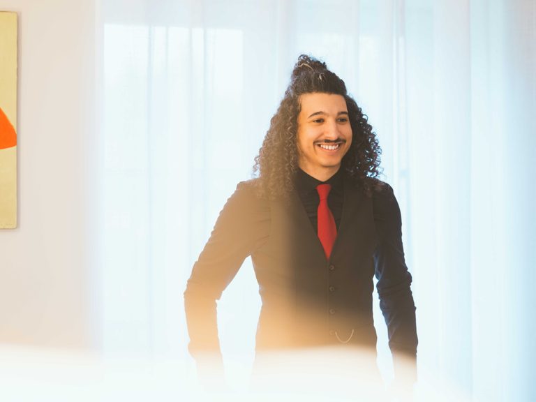 Marouane souriant en gilet noir et cravate rouge devant les voilages, préparatifs au Château d'Auvilliers