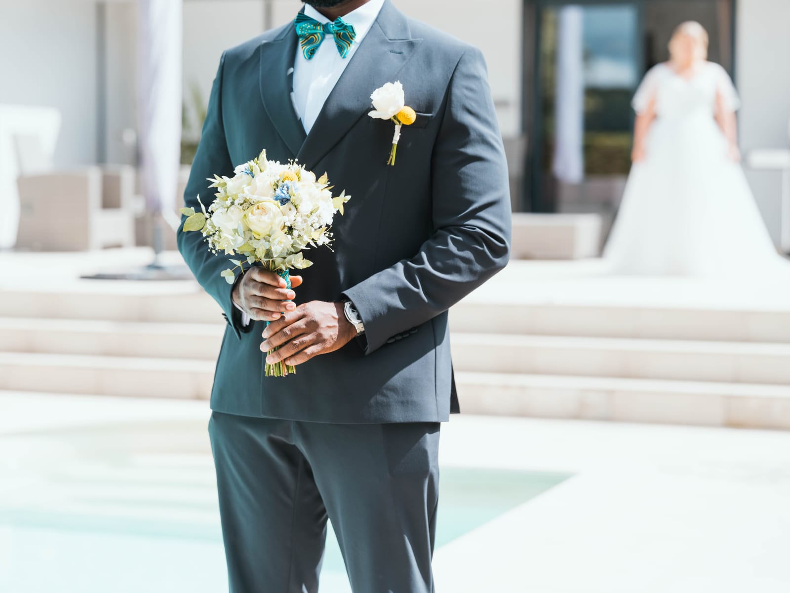Issa descendant les marches du perron du château avec le bouquet, costume gris, façade classique en arrière-plan, mariage au Domaine du Mont Rouge, par Jean-Baptiste Argyre (Jaysphotography)