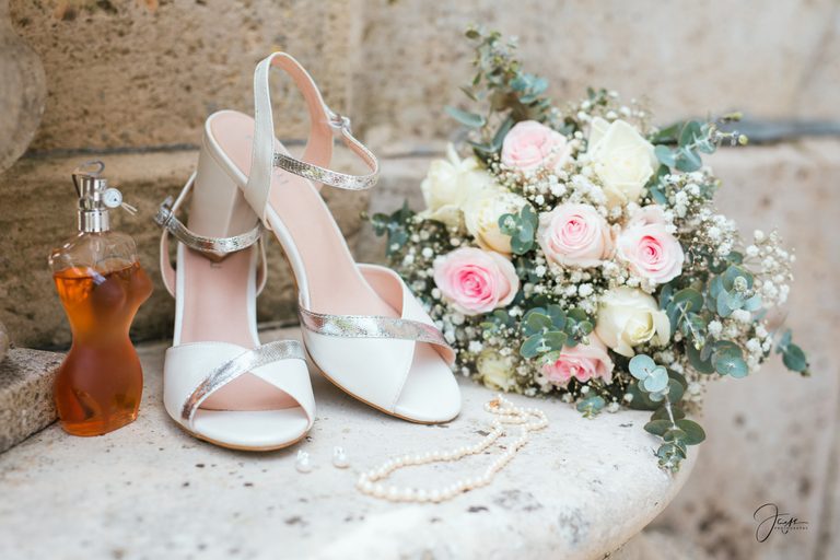 Sandales blanches de Doralie, bouquet de roses pastel, parfum et collier de perles posés sur la pierre, préparatifs au Château de Broyes