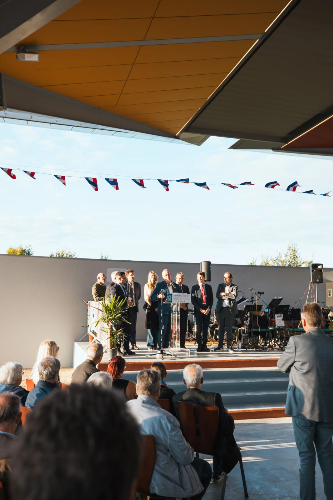 La tribune officielle vue depuis le public assis, sous les ombrelles ocre de la halle couverte de Breuil-le-Vert, mariage au Breuil-le-Vert (Oise), par Jean-Baptiste Argyre (Jaysphotography)