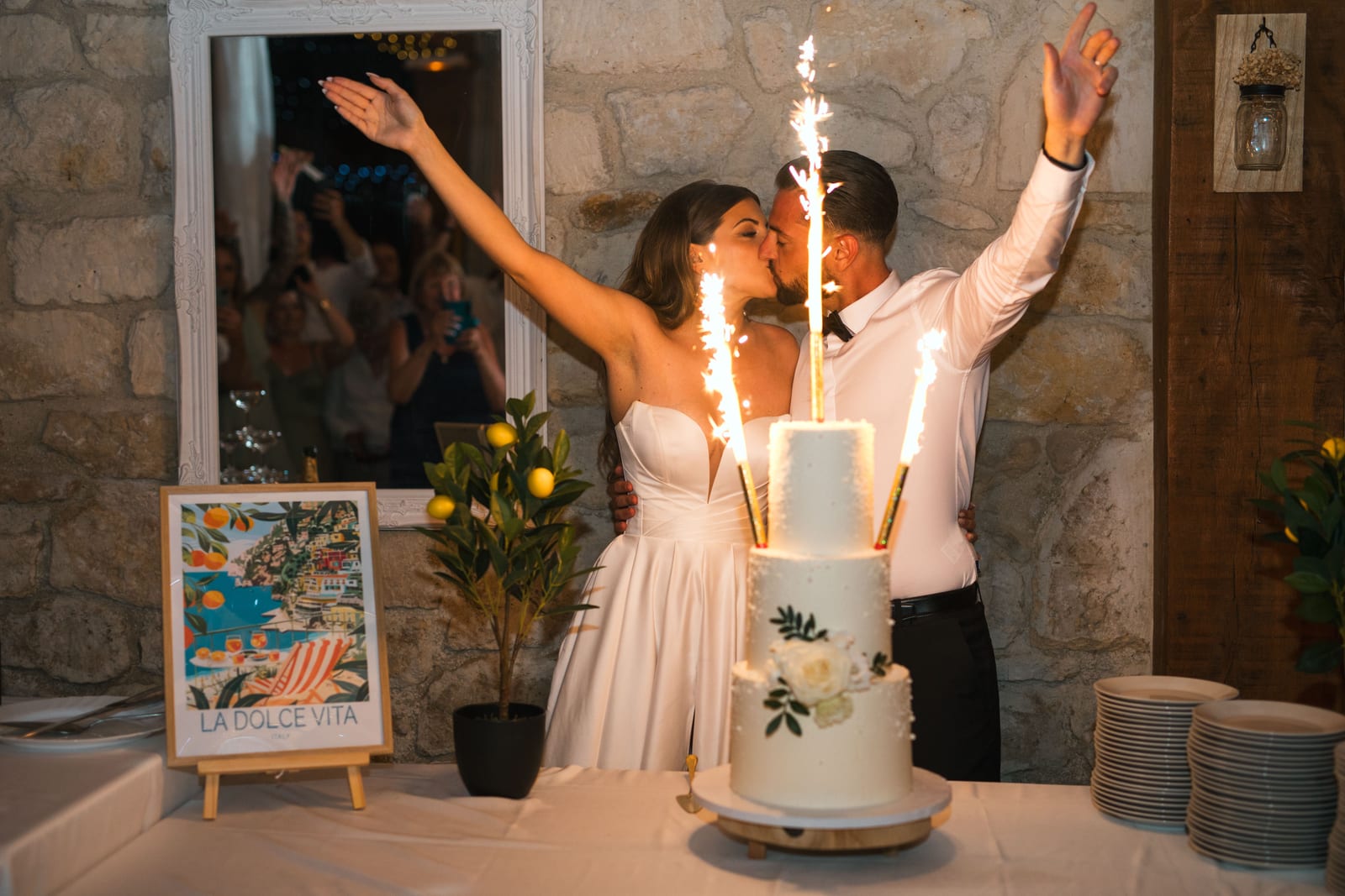 Killian et Élodie devant le gâteau blanc trois étages avec l'affiche Dolce Vita, bras levés, Domaine de l'Orangerie, mariage au Domaine de l'Orangerie, par Jean-Baptiste Argyre (Jaysphotography)