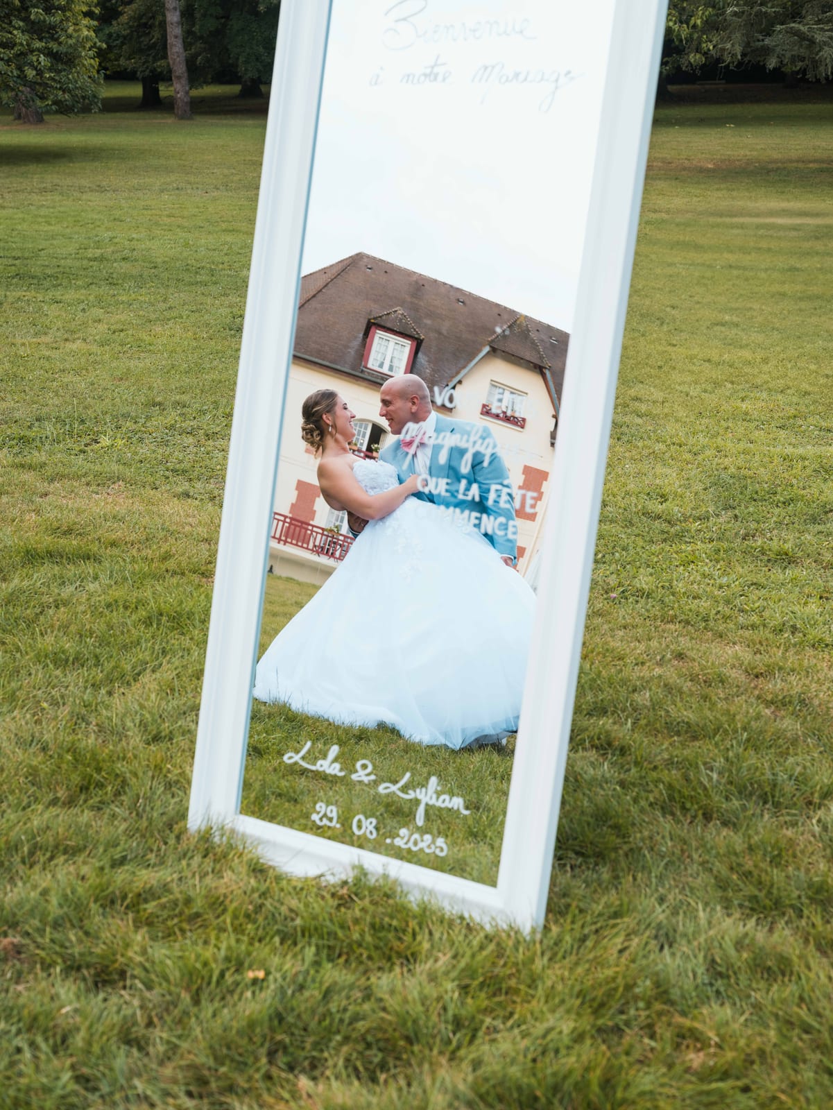 Miroir personnalisé avec les prénoms Lola et Lylian et la date 29.08.2025 installé dans l'herbe verte du parc, façade normande reflétée dans la glace, mariage au Château de La Tour, par Jean-Baptiste Argyre (Jaysphotography)