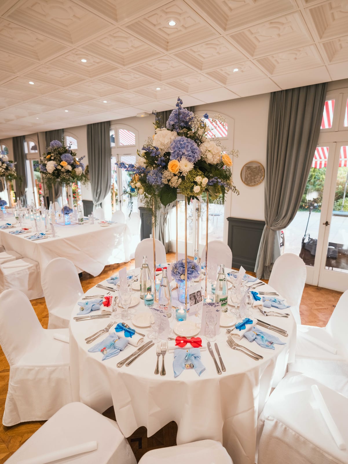 Salle de réception du château décorée pour le dîner, tables rondes dressées avec centres de table fleuris blanc et bleu, guirlandes lumineuses, mariage au Château de La Tour, par Jean-Baptiste Argyre (Jaysphotography)