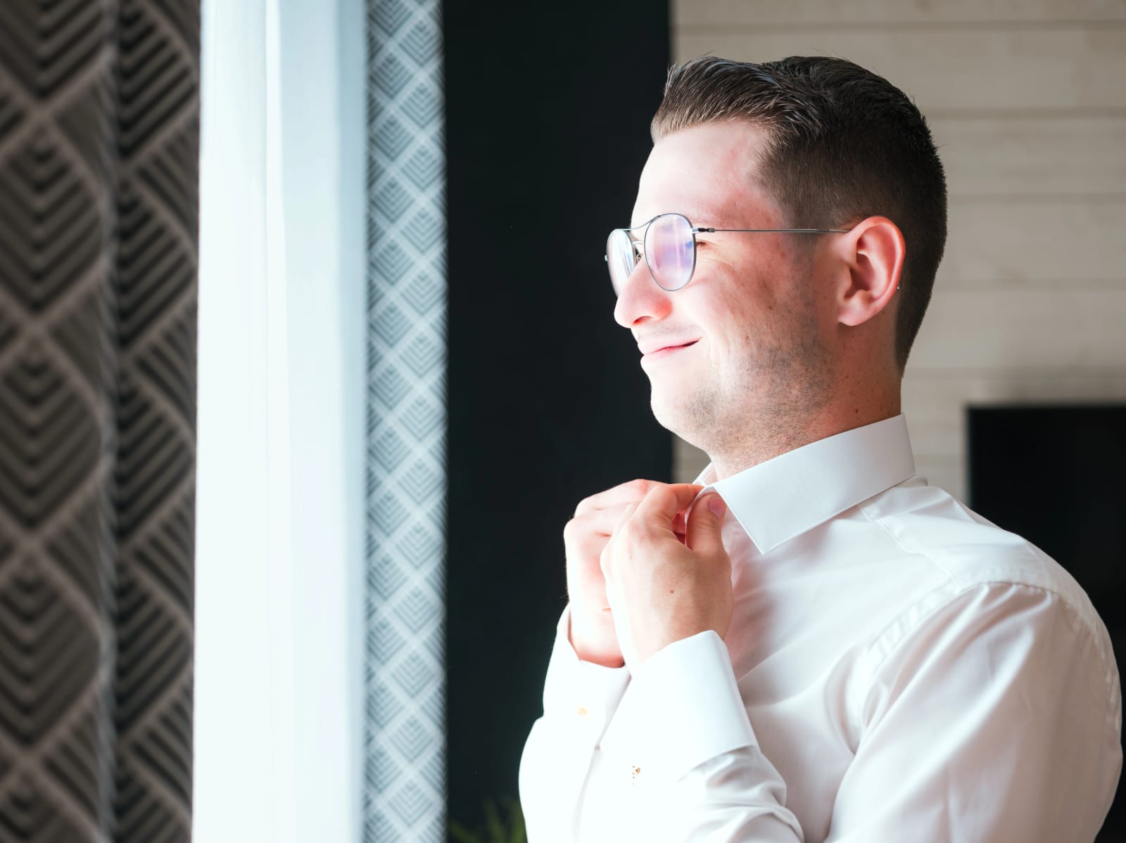 Alex de profil ajuste sa cravate devant la fenêtre, lunettes rondes, dans la lumière des préparatifs, mariage au Domaine du Bois Saint Mard, par Jean-Baptiste Argyre (Jaysphotography)