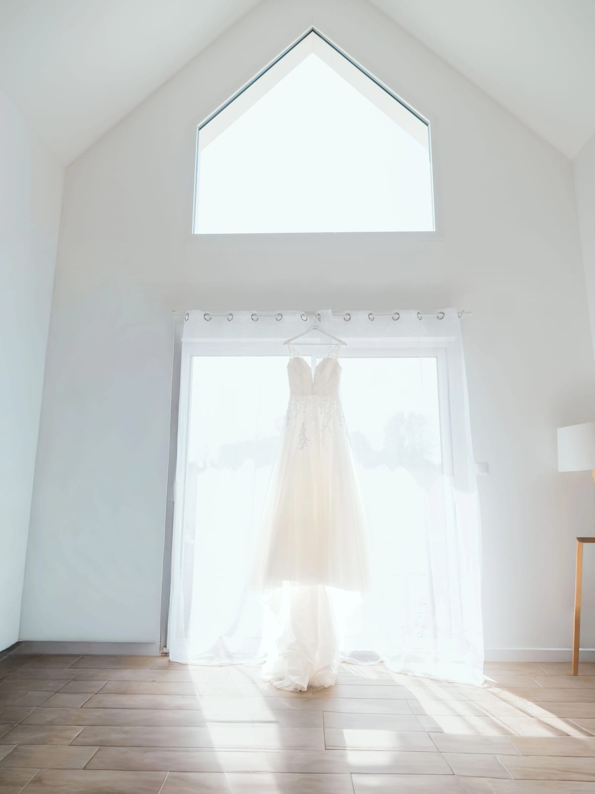 La robe de mariée ivoire en dentelle suspendue devant une grande fenêtre claire avant l'habillage, mariage au Domaine du Bois Saint Mard, par Jean-Baptiste Argyre (Jaysphotography)