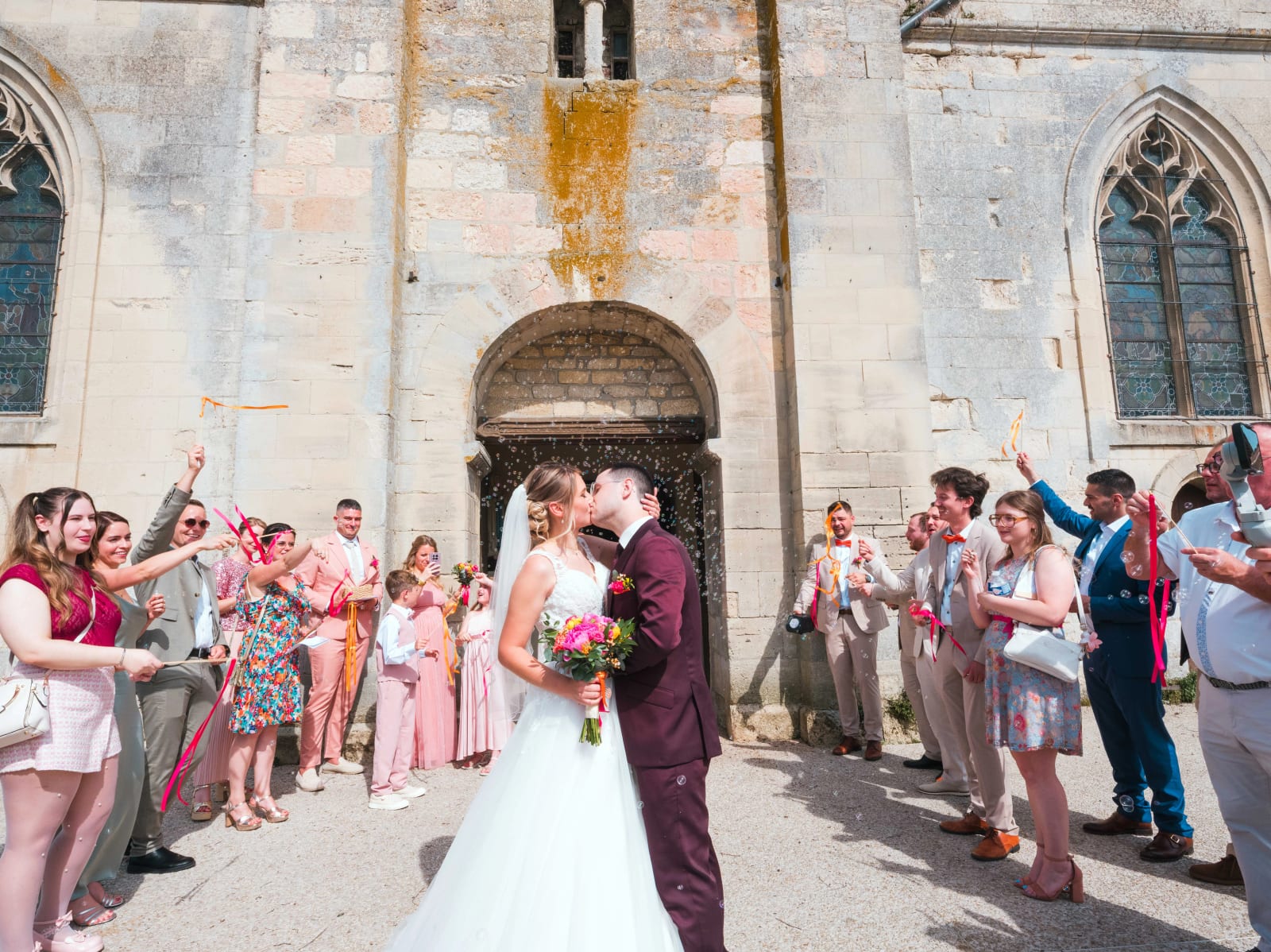 Les mariés s'embrassent à la sortie de l'église sous une haie de rubans colorés et de bulles, mariage au Domaine du Bois Saint Mard, par Jean-Baptiste Argyre (Jaysphotography)