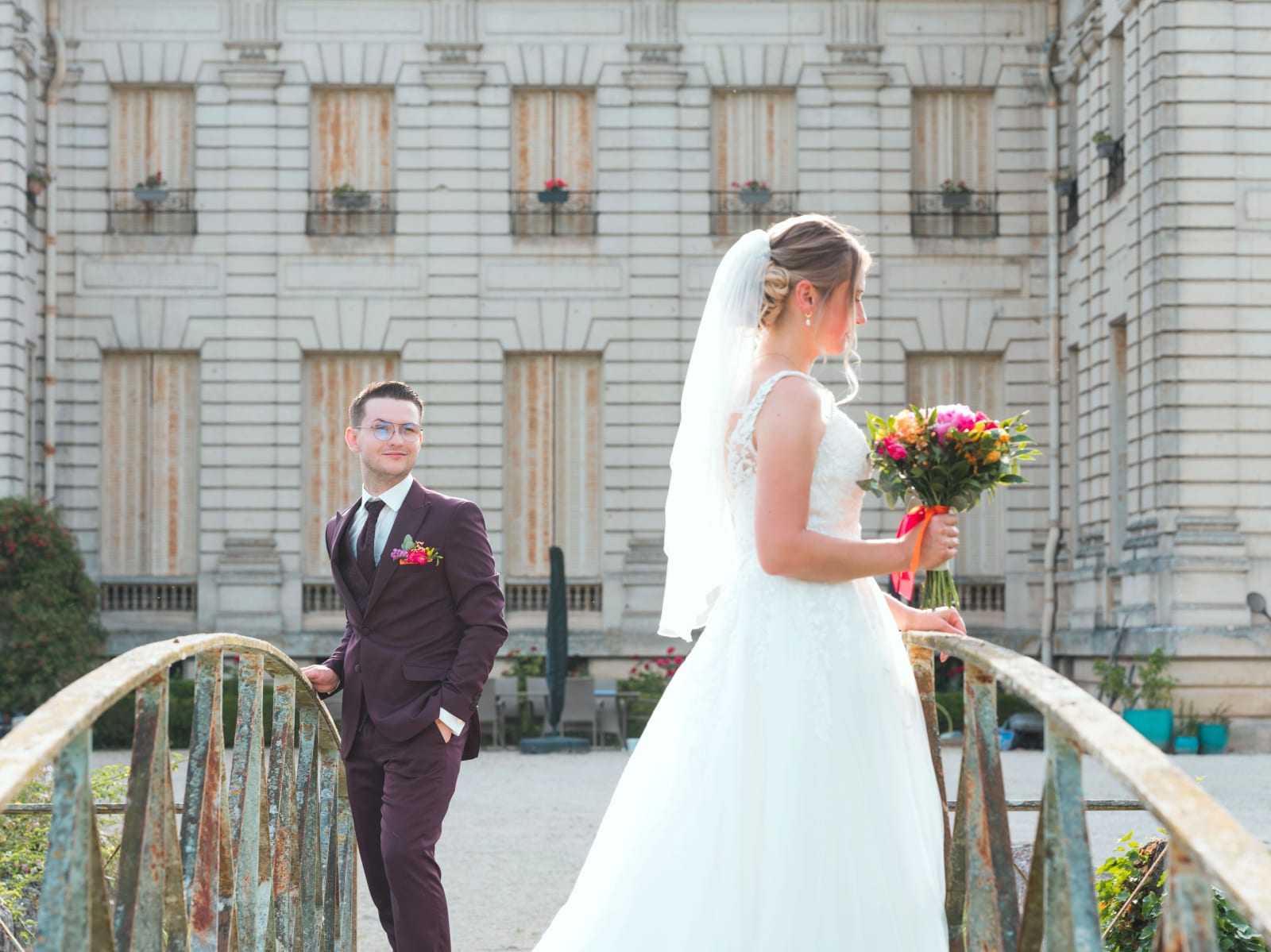 Alex regarde Audrey sur le vieux pont métallique du domaine, son bouquet coloré à la main, mariage au Domaine du Bois Saint Mard, par Jean-Baptiste Argyre (Jaysphotography)