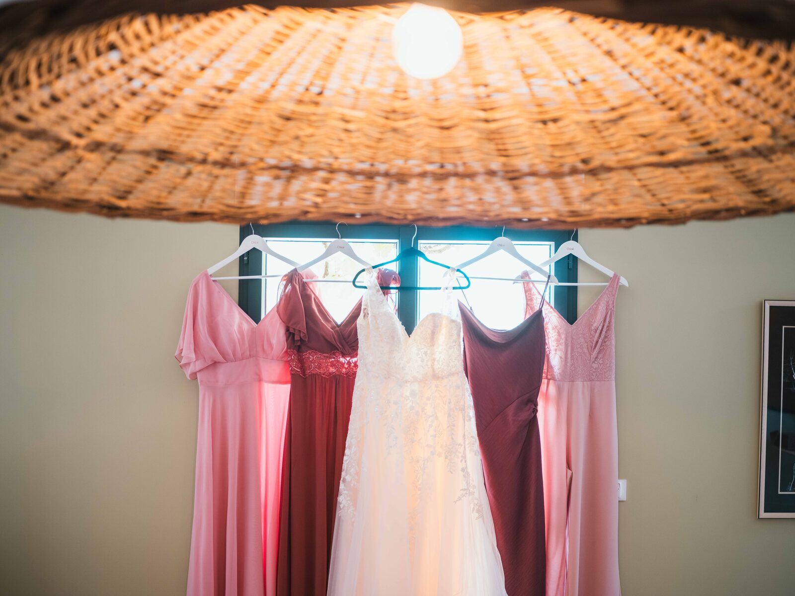 Les robes des demoiselles d'honneur en dégradé de rose et la robe de mariée en dentelle suspendues sous un grand luminaire en osier — mariage au Domaine du Bois Saint Mard, par Jean-Baptiste Argyre (Jaysphotography)