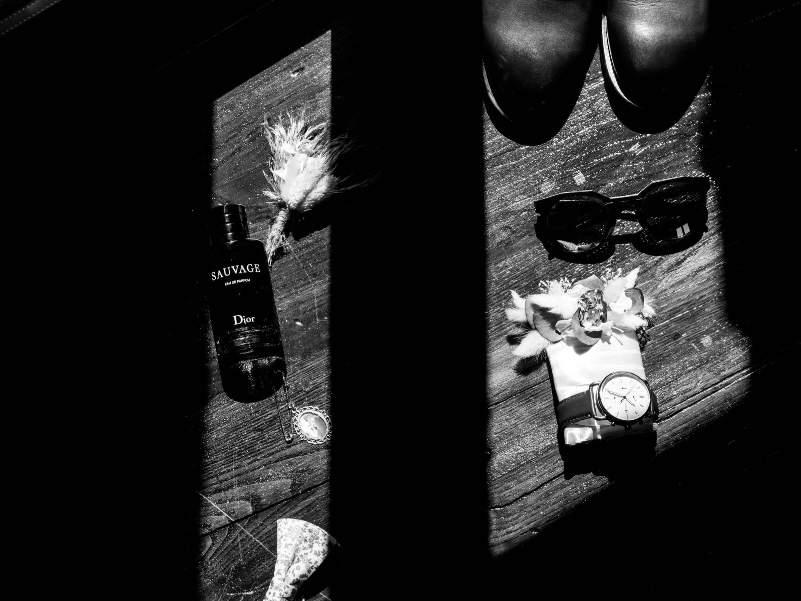 Détail noir et blanc des accessoires du marié : parfum, montre, lunettes de soleil et boutonnière posés sur le parquet — mariage au Domaine du Bois Saint Mard, par Jean-Baptiste Argyre (Jaysphotography)
