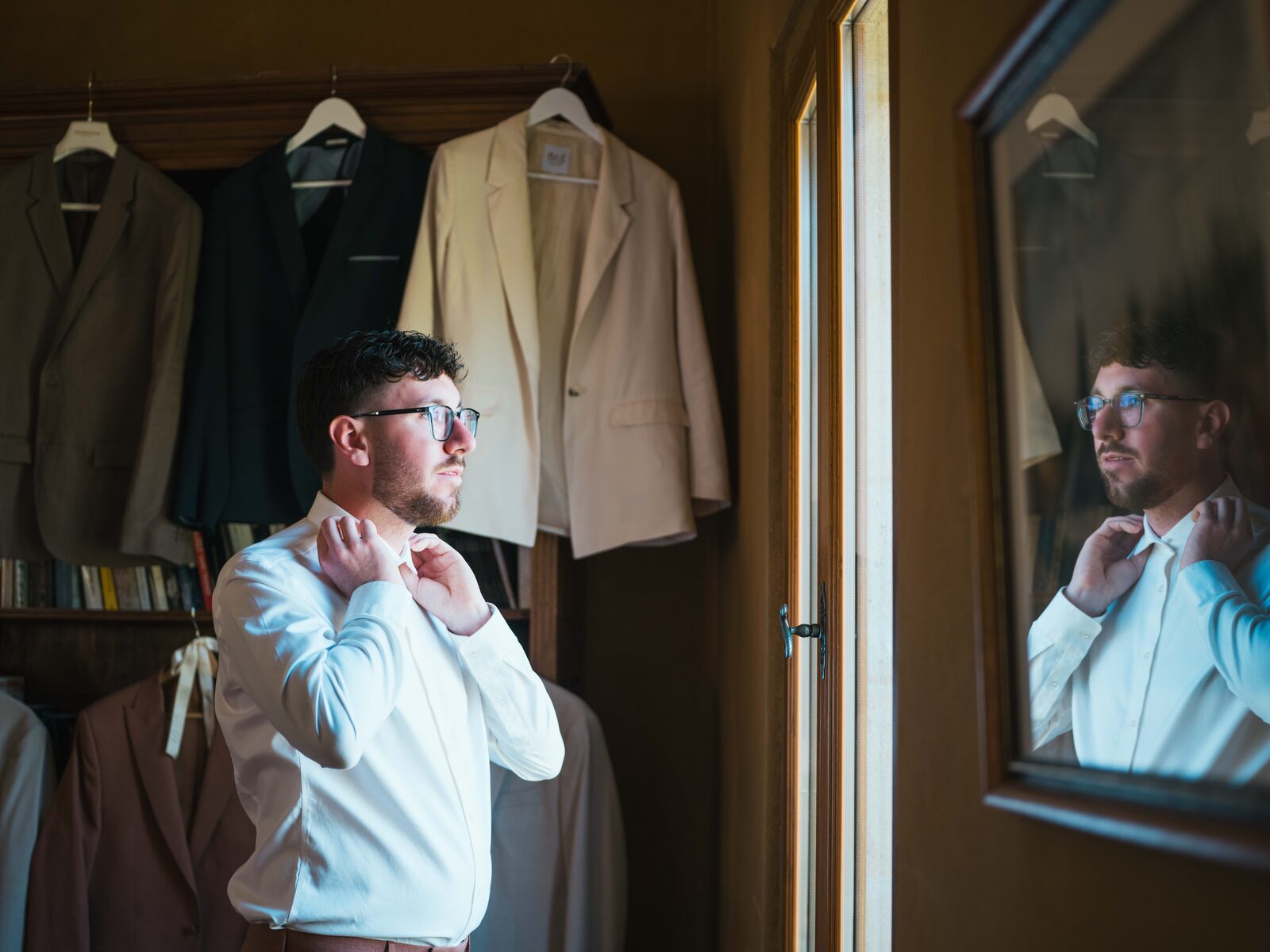 Le marié en chemise blanche ajuste son col devant le miroir, ses costumes suspendus derrière lui — mariage au Domaine du Bois Saint Mard, par Jean-Baptiste Argyre (Jaysphotography)