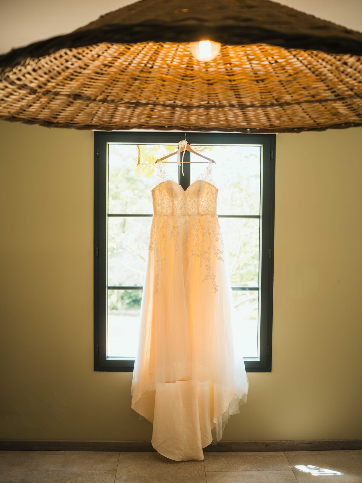 La robe de mariée en dentelle suspendue à la fenêtre, baignée de lumière — mariage au Domaine du Bois Saint Mard, par Jean-Baptiste Argyre (Jaysphotography)