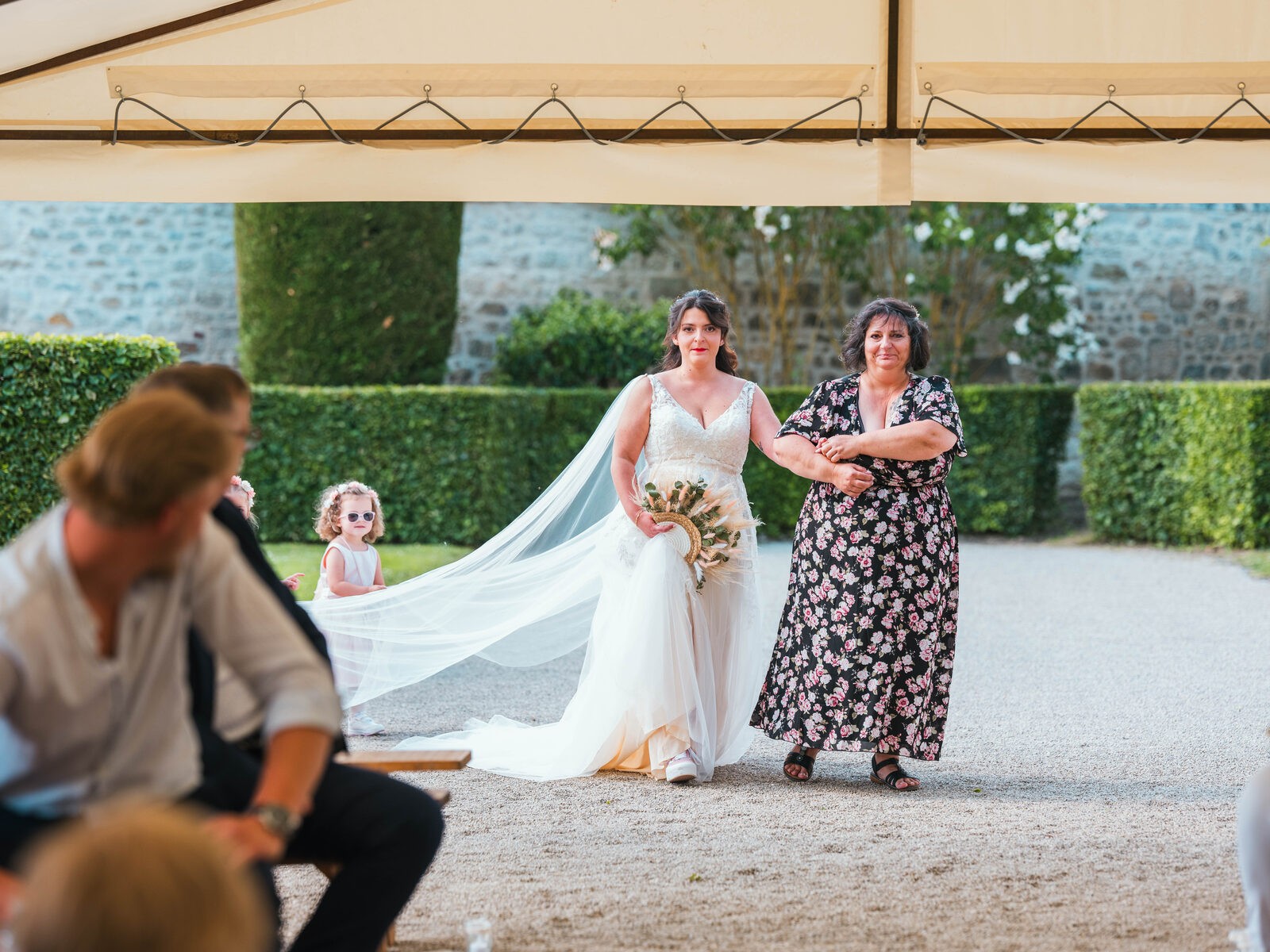 Entrée de la mariée au bras de sa maman, longue traîne et voile, sous le regard d'une petite invitée — mariage au Domaine du Bois Saint Mard, par Jean-Baptiste Argyre (Jaysphotography)