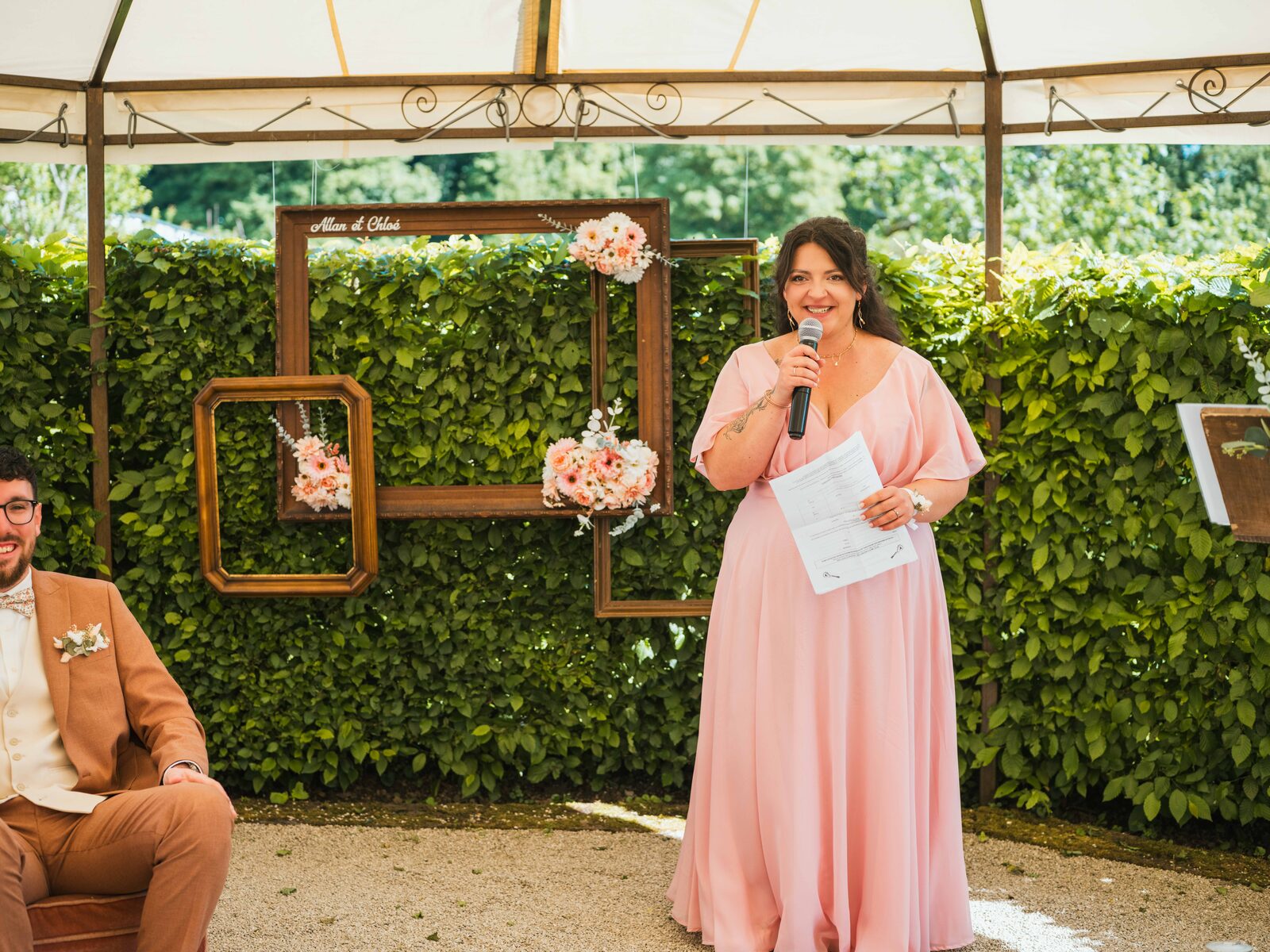 Une oratrice en robe rose prend la parole devant le décor de cadres « Allan & Chloé », le marié assis à ses côtés — mariage au Domaine du Bois Saint Mard, par Jean-Baptiste Argyre (Jaysphotography)