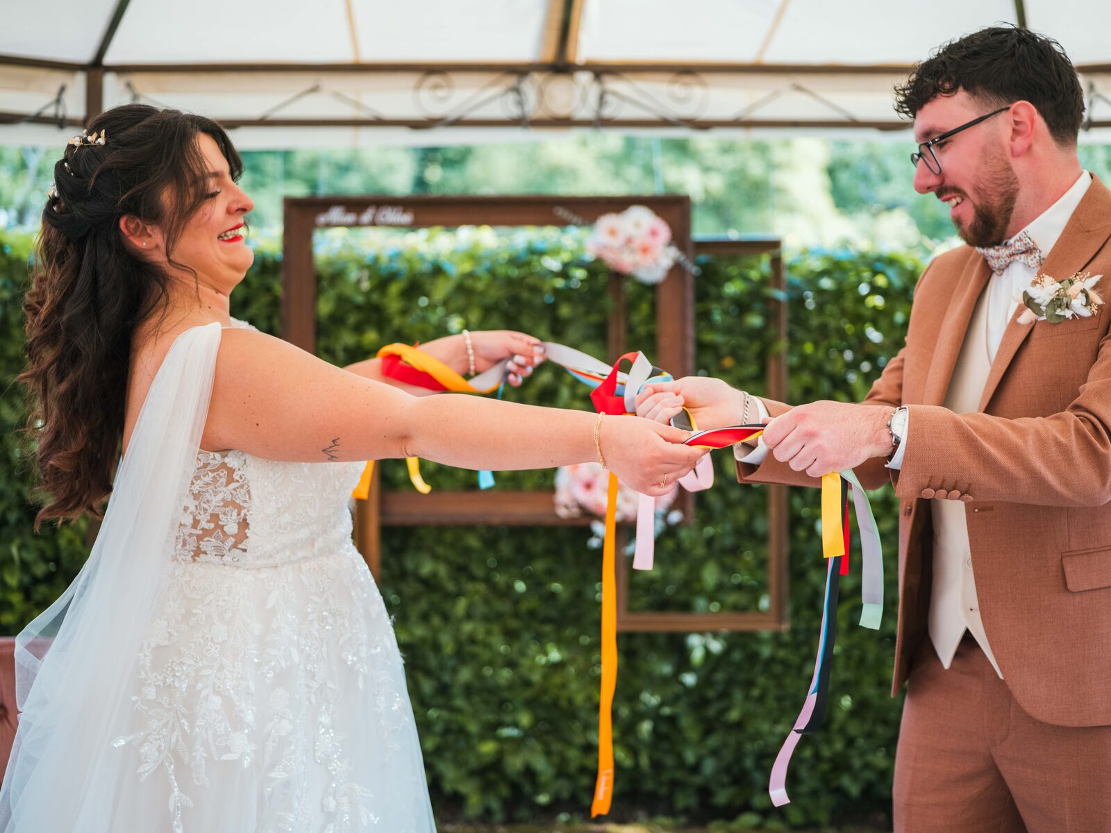 Les mariés rient face à face en tirant sur les rubans colorés du rituel pendant la cérémonie — mariage au Domaine du Bois Saint Mard, par Jean-Baptiste Argyre (Jaysphotography)
