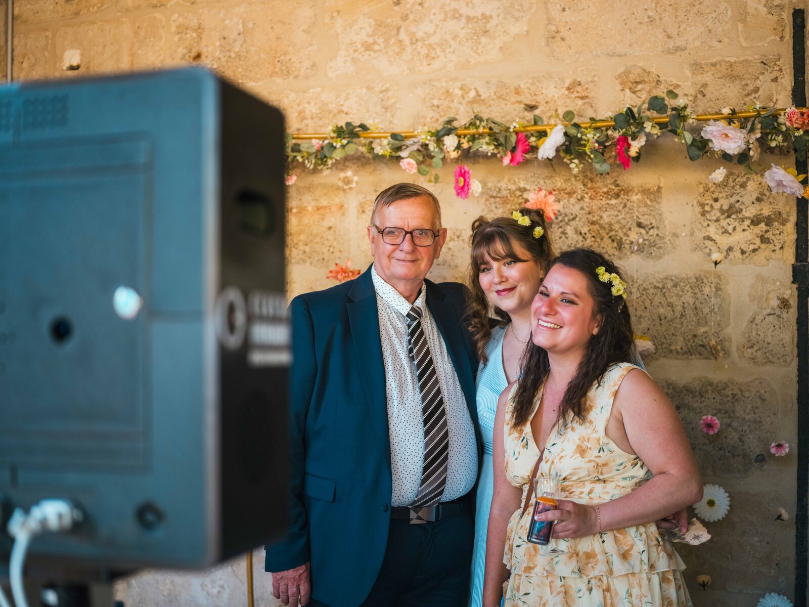 Un grand-père et deux jeunes femmes posent devant le photobooth et son mur fleuri — mariage au Domaine du Bois Saint Mard, par Jean-Baptiste Argyre (Jaysphotography)