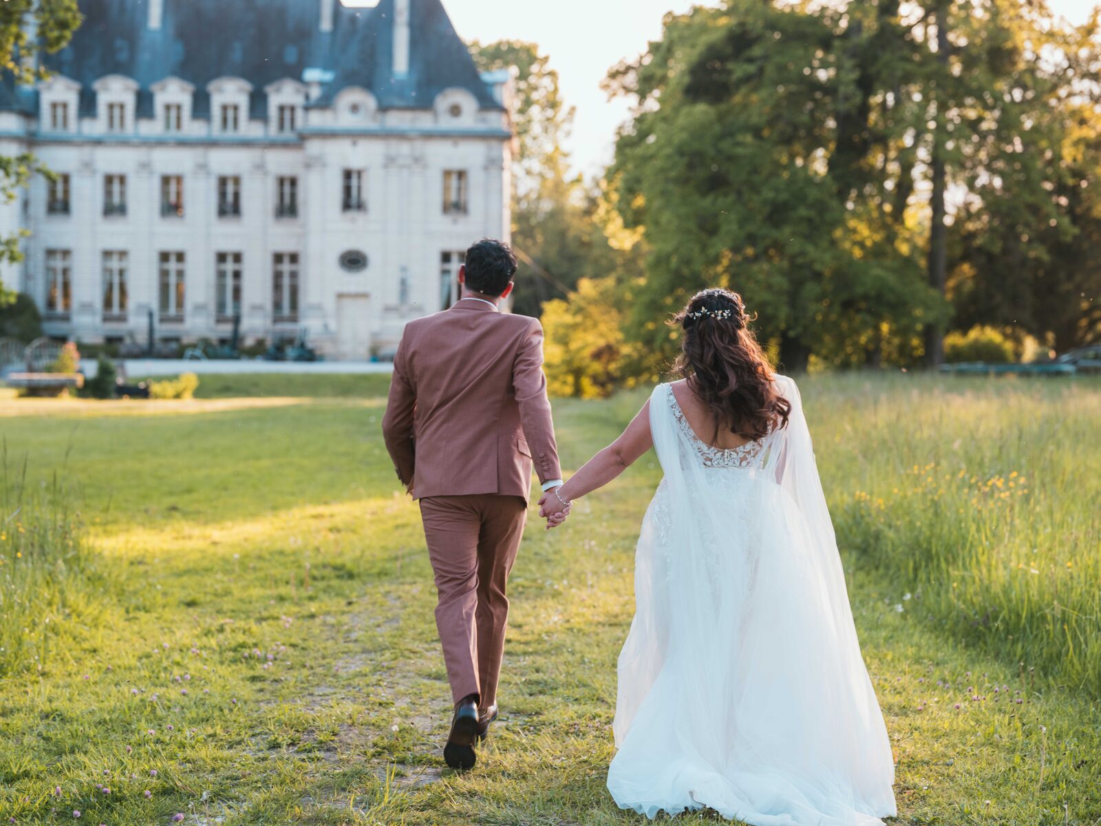 Les mariés de dos marchent vers le château dans la prairie dorée du soir — mariage au Domaine du Bois Saint Mard, par Jean-Baptiste Argyre (Jaysphotography)