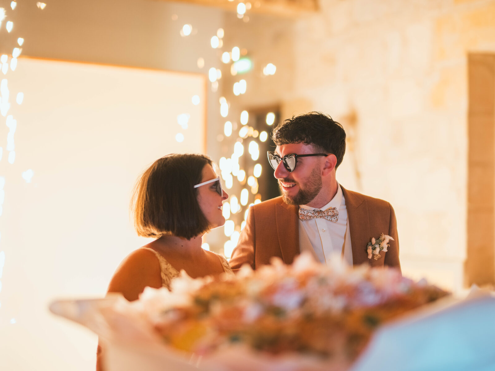 Les mariés complices devant un rideau de guirlandes lumineuses, le dessert au premier plan — mariage au Domaine du Bois Saint Mard, par Jean-Baptiste Argyre (Jaysphotography)