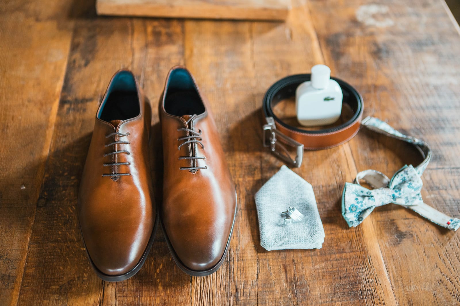 Flat lay des accessoires du marié : chaussures richelieu cognac, ceinture en cuir, nœud papillon liberty fleuri, parfum et boutons de manchette sur une table en bois, mariage au Domaine de Senercy, par Jean-Baptiste Argyre (Jaysphotography)