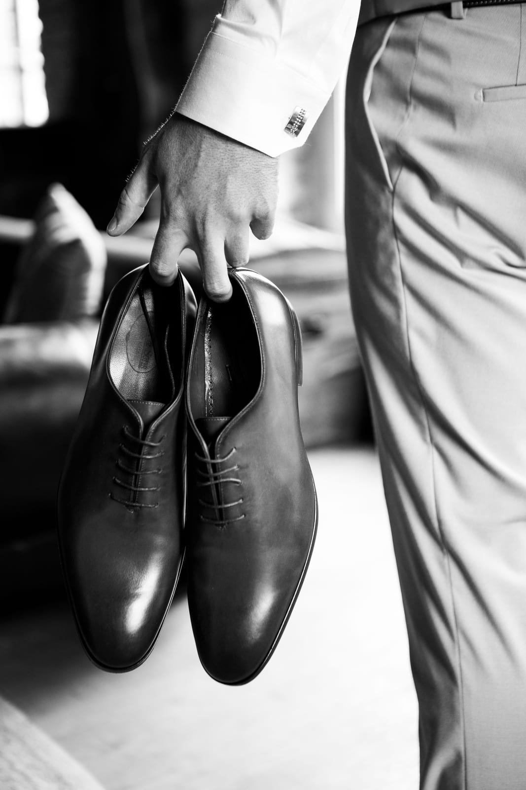 Benjamin en pantalon de costume gris tient ses chaussures de ville en cuir à la main, photo en noir et blanc pendant l'habillage, mariage au Domaine de Senercy, par Jean-Baptiste Argyre (Jaysphotography)