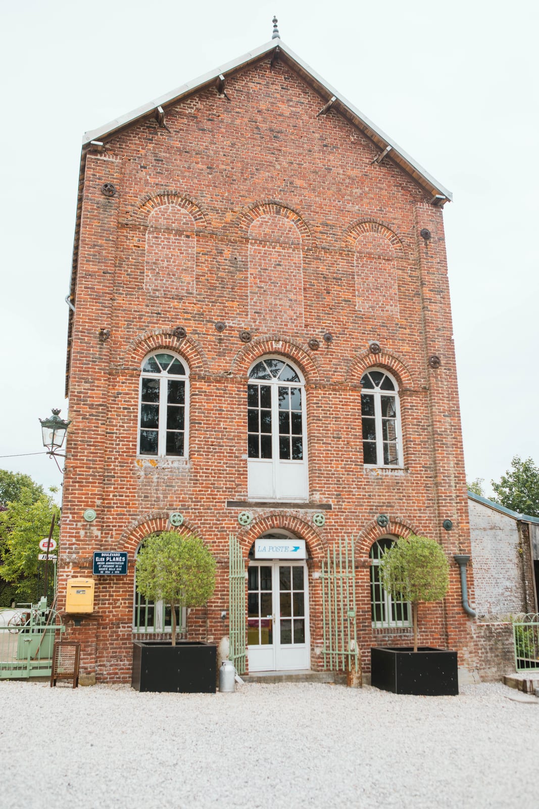 Façade en brique rouge d'un ancien bâtiment de poste du Domaine de Senercy, allée gravillonnée et arbustes taillés, mariage au Domaine de Senercy, par Jean-Baptiste Argyre (Jaysphotography)