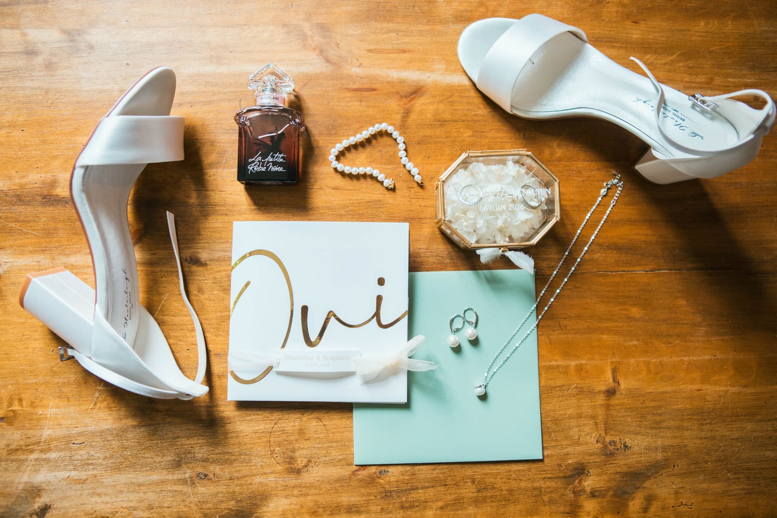Flat lay des accessoires d'Amandine : sandales blanches à talon, parfum, collier de perles, alliances et faire-part doré "Oui", mariage au Domaine de Senercy, par Jean-Baptiste Argyre (Jaysphotography)