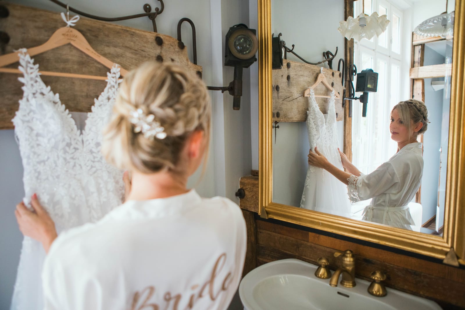 Amandine en peignoir tient sa robe de mariée devant le grand miroir doré et la robinetterie en laiton du Domaine de Senercy, mariage au Domaine de Senercy, par Jean-Baptiste Argyre (Jaysphotography)