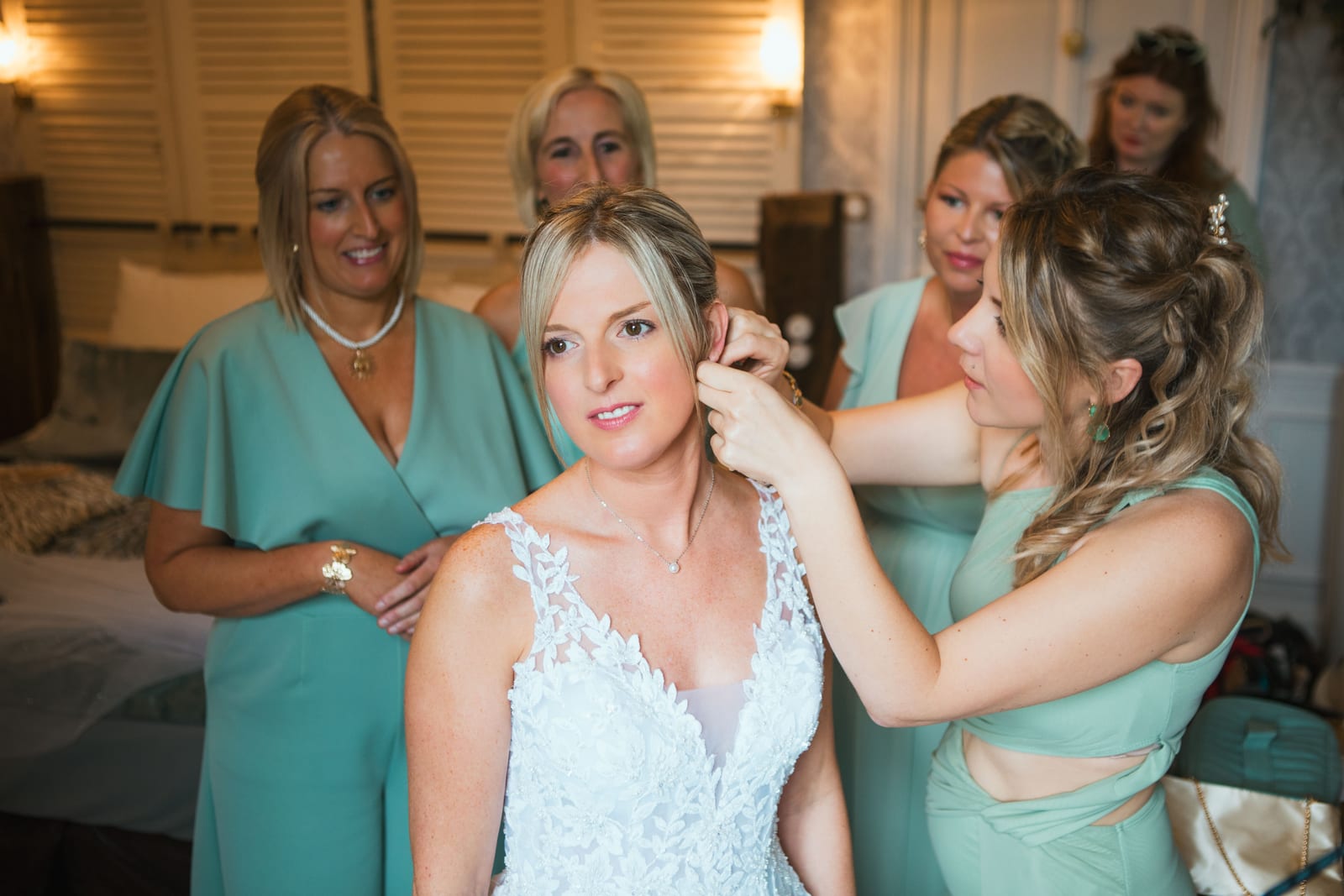 Une demoiselle d'honneur en robe vert d'eau attache la boucle d'oreille d'Amandine en robe de dentelle, entourée de ses proches, mariage au Domaine de Senercy, par Jean-Baptiste Argyre (Jaysphotography)