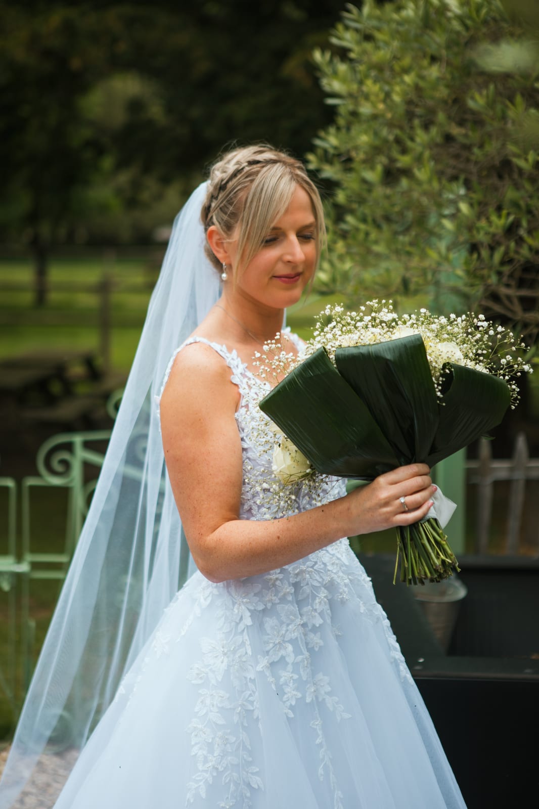 Amandine de profil regarde son bouquet de roses blanches et de gypsophile, voile au vent, dans le parc du domaine, mariage au Domaine de Senercy, par Jean-Baptiste Argyre (Jaysphotography)