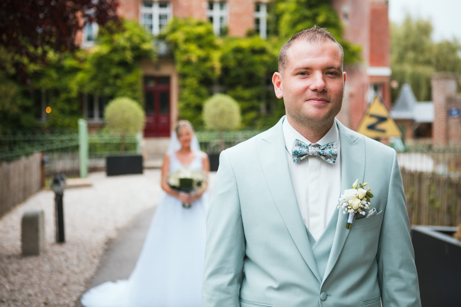 First look : Benjamin en costume vert d'eau au premier plan tandis qu'Amandine s'avance derrière lui dans l'allée du domaine, mariage au Domaine de Senercy, par Jean-Baptiste Argyre (Jaysphotography)
