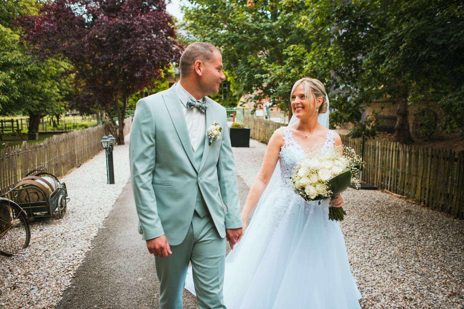 Amandine et Benjamin marchent main dans la main dans l'allée gravillonnée du domaine en se souriant, mariage au Domaine de Senercy, par Jean-Baptiste Argyre (Jaysphotography)