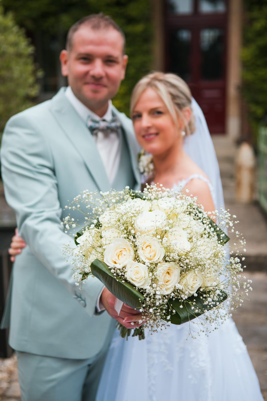 Le bouquet de roses blanches et de gypsophile au premier plan, Amandine et Benjamin enlacés en arrière-plan devant une porte rouge, mariage au Domaine de Senercy, par Jean-Baptiste Argyre (Jaysphotography)