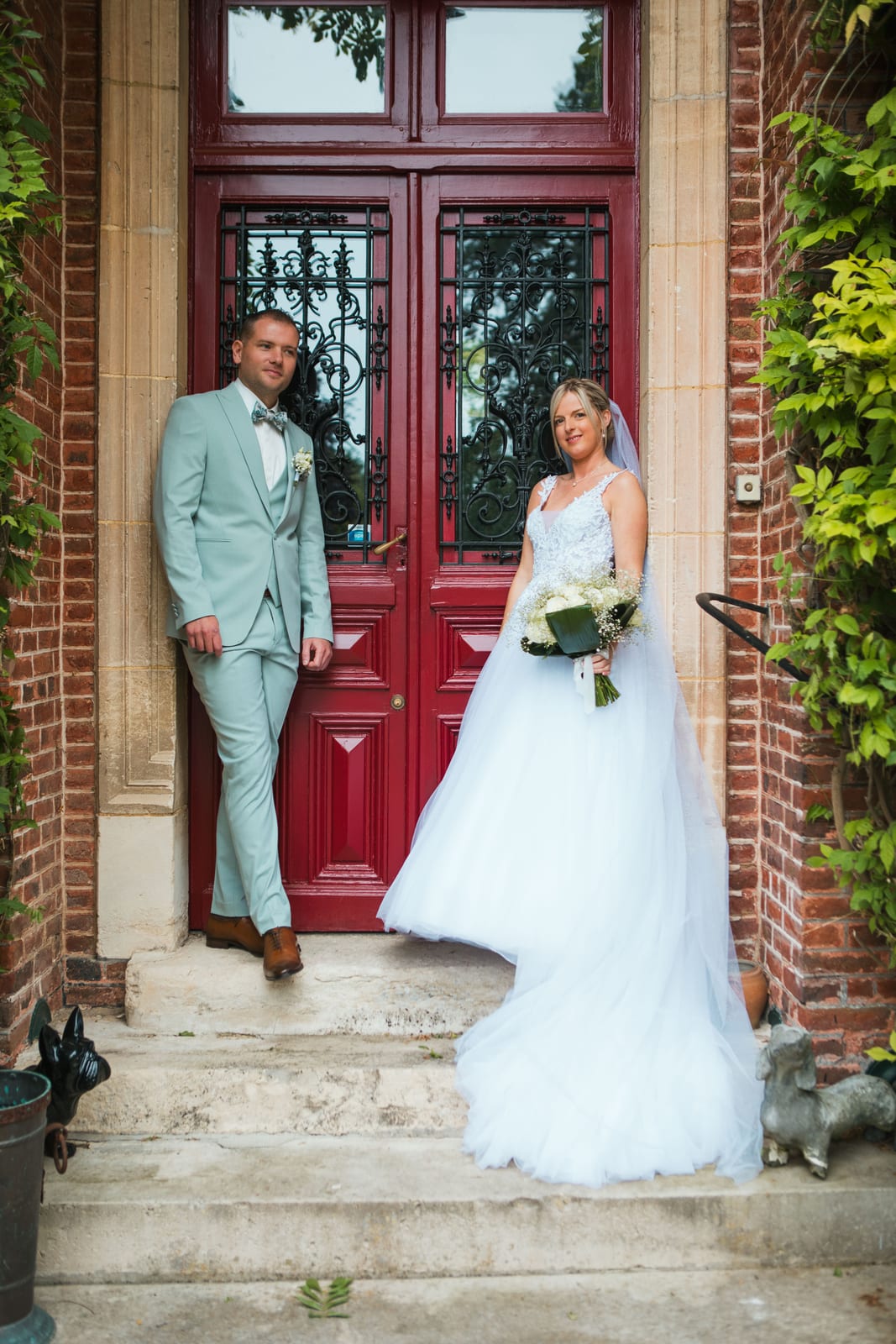 Amandine et Benjamin posent dans l'encadrement de la grande porte rouge à ferronnerie du manoir en brique, mariage au Domaine de Senercy, par Jean-Baptiste Argyre (Jaysphotography)