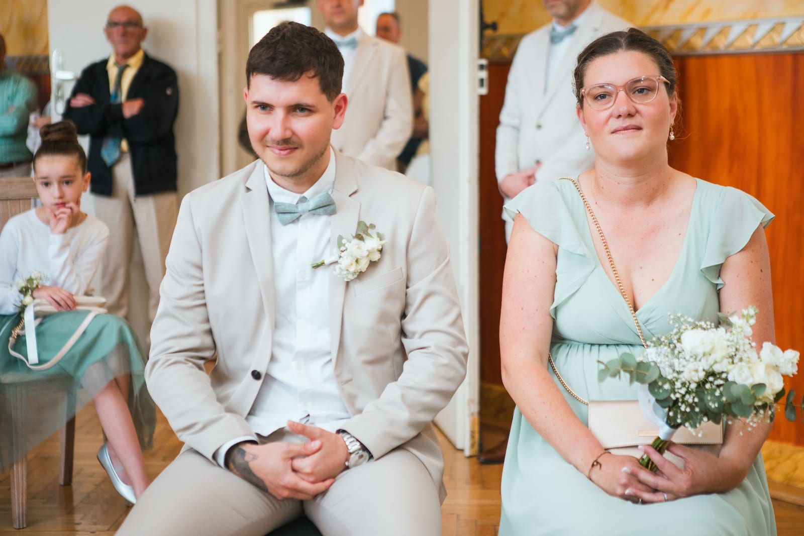 Le témoin en costume beige et la témoin en robe vert d'eau suivent la cérémonie civile au premier rang, une enfant d'honneur derrière, mariage au Domaine de Senercy, par Jean-Baptiste Argyre (Jaysphotography)