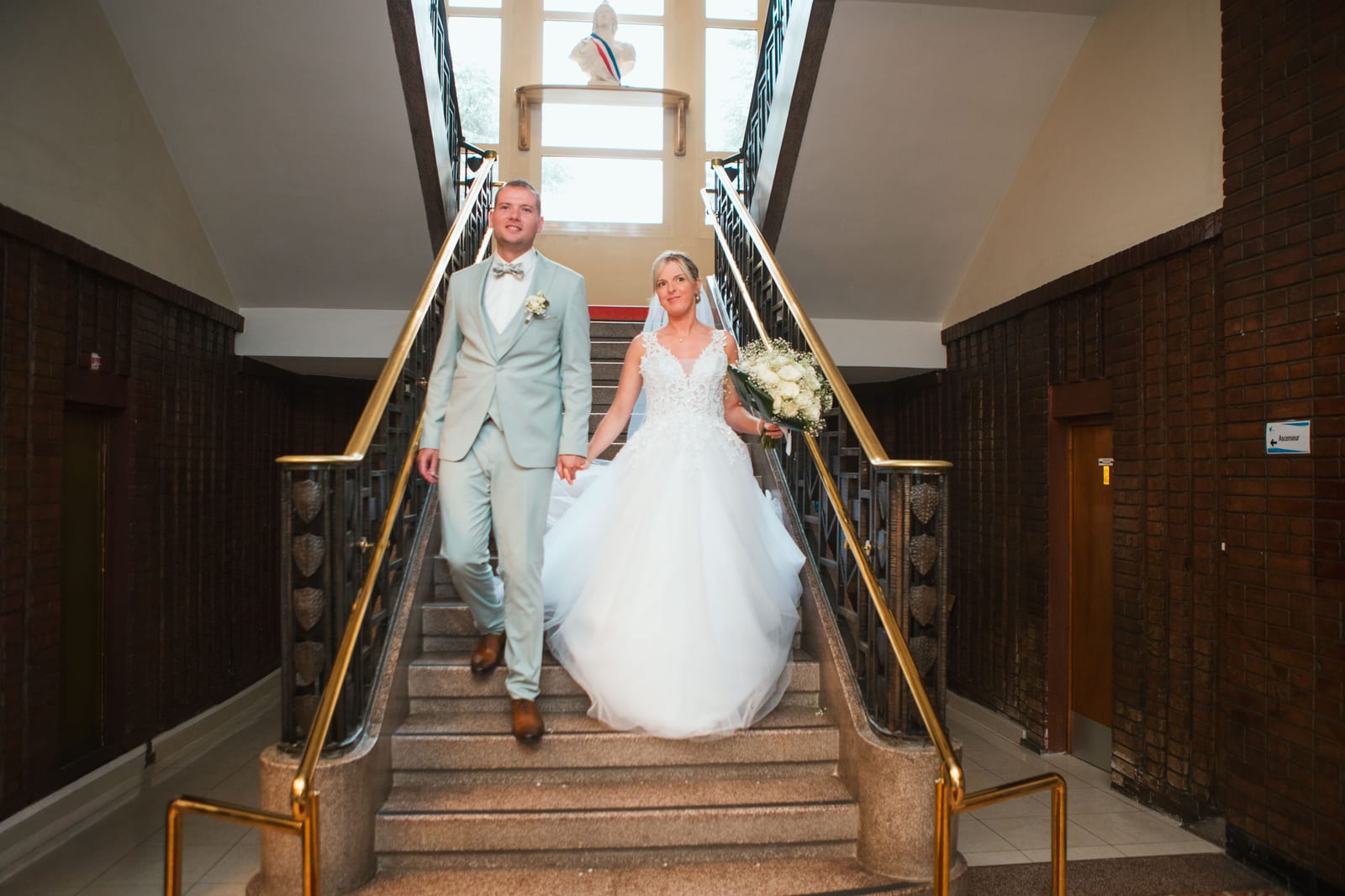 Amandine et Benjamin descendent main dans la main le grand escalier en laiton de la mairie sous le buste de Marianne, mariage au Domaine de Senercy, par Jean-Baptiste Argyre (Jaysphotography)