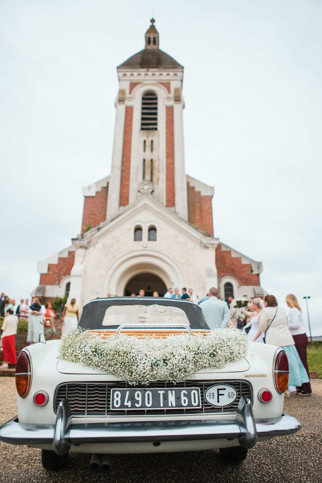 La voiture de collection blanche décorée d'une guirlande de gypsophile devant l'église de Tergnier, invités sur le parvis, mariage au Domaine de Senercy, par Jean-Baptiste Argyre (Jaysphotography)