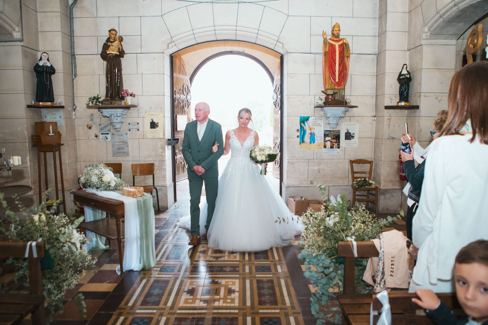 Amandine entre dans l'église de Tergnier au bras de son père, à contre-jour dans le portail, allée fleurie d'eucalyptus, mariage au Domaine de Senercy, par Jean-Baptiste Argyre (Jaysphotography)