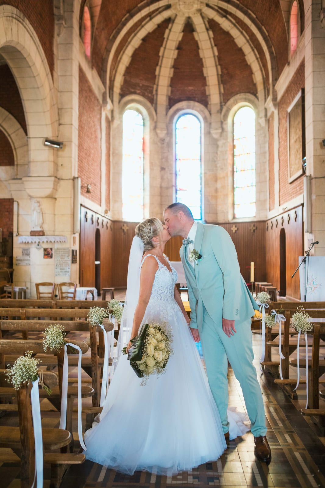 Amandine et Benjamin s'embrassent dans l'allée de l'église de Tergnier aux voûtes de brique, bancs ornés de gypsophile, mariage au Domaine de Senercy, par Jean-Baptiste Argyre (Jaysphotography)
