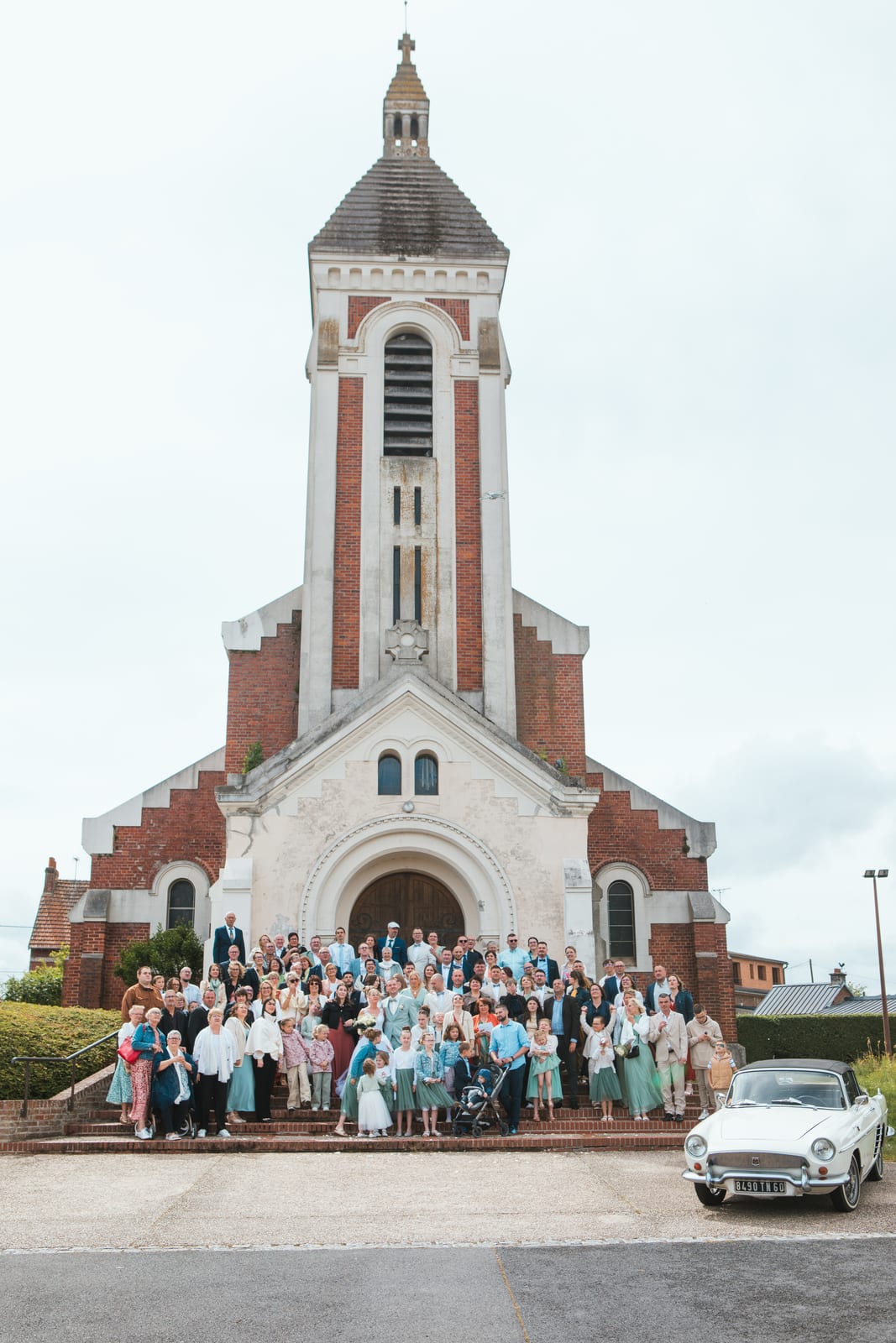 Photo de groupe des invités sur les marches de l'église de Tergnier, la voiture de collection blanche à droite, mariage au Domaine de Senercy, par Jean-Baptiste Argyre (Jaysphotography)
