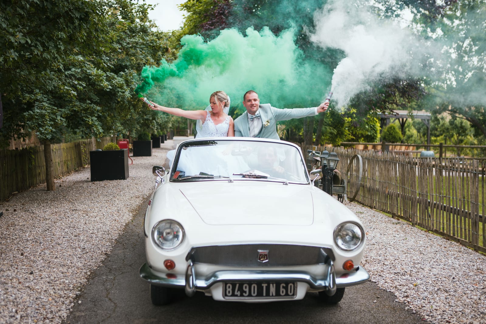 Amandine et Benjamin debout dans la décapotable blanche brandissent des fumigènes verts dans l'allée du Domaine de Senercy, mariage au Domaine de Senercy, par Jean-Baptiste Argyre (Jaysphotography)