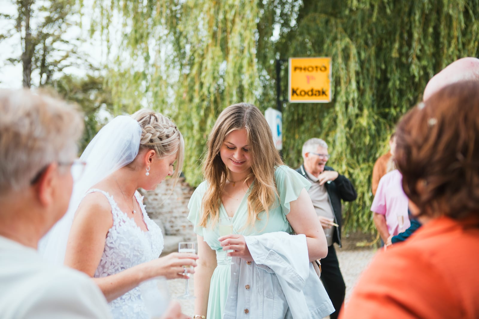 Amandine discute avec une invitée, coupe à la main, au vin d'honneur près d'un panneau émaillé vintage "Photo Kodak", mariage au Domaine de Senercy, par Jean-Baptiste Argyre (Jaysphotography)