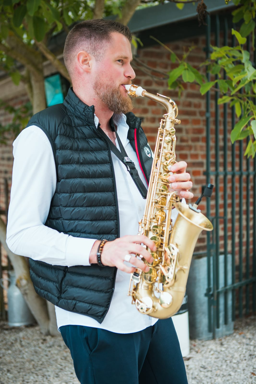 Le saxophoniste, doudoune noire et saxophone doré, joue en live pendant le vin d'honneur dans la cour du domaine, mariage au Domaine de Senercy, par Jean-Baptiste Argyre (Jaysphotography)
