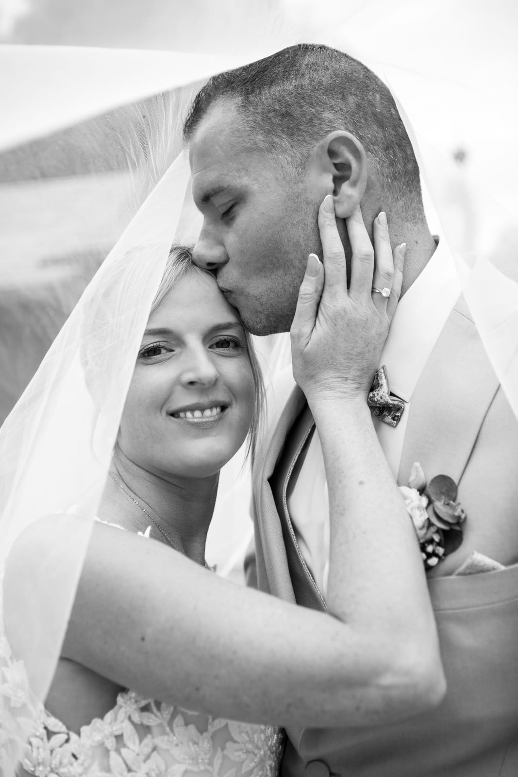 Portrait de couple en noir et blanc sous le voile : Benjamin embrasse le front d'Amandine qui sourit, mariage au Domaine de Senercy, par Jean-Baptiste Argyre (Jaysphotography)