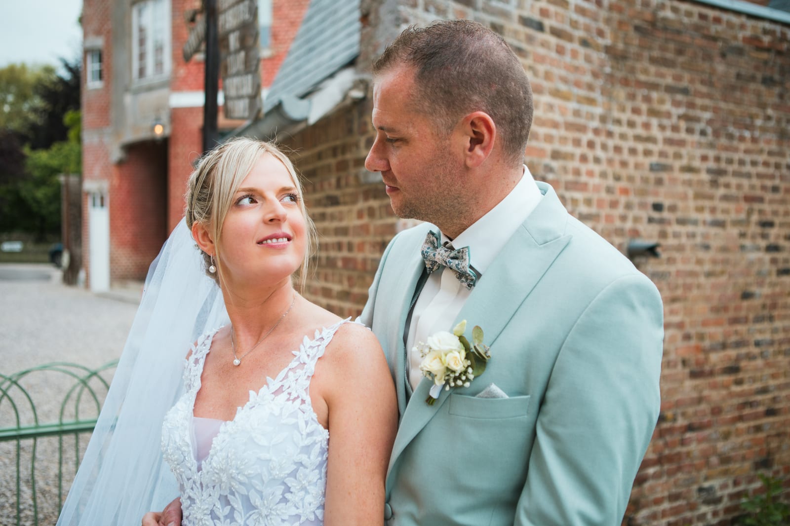 Amandine lève les yeux vers Benjamin en costume vert d'eau devant un mur de brique du Domaine de Senercy, mariage au Domaine de Senercy, par Jean-Baptiste Argyre (Jaysphotography)