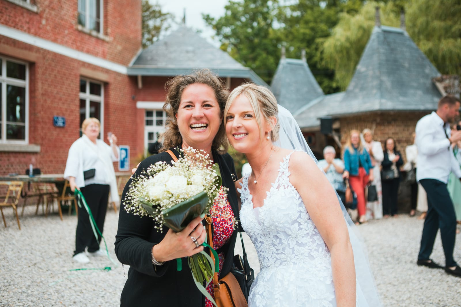 Amandine, joue contre joue avec une proche tenant le bouquet, rit devant le manoir aux toitures en poivrière, mariage au Domaine de Senercy, par Jean-Baptiste Argyre (Jaysphotography)