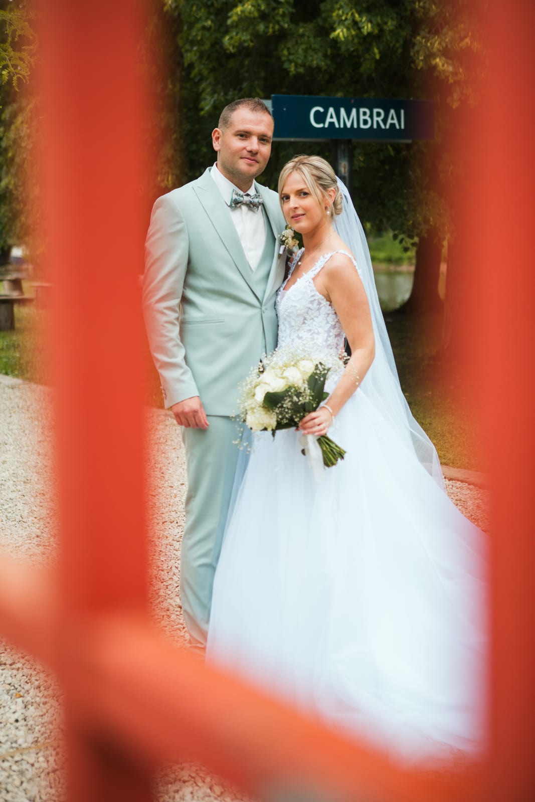 Amandine et Benjamin posent près de l'étang, cadrés à travers une structure rouge, sous un panneau vintage "Cambrai", mariage au Domaine de Senercy, par Jean-Baptiste Argyre (Jaysphotography)