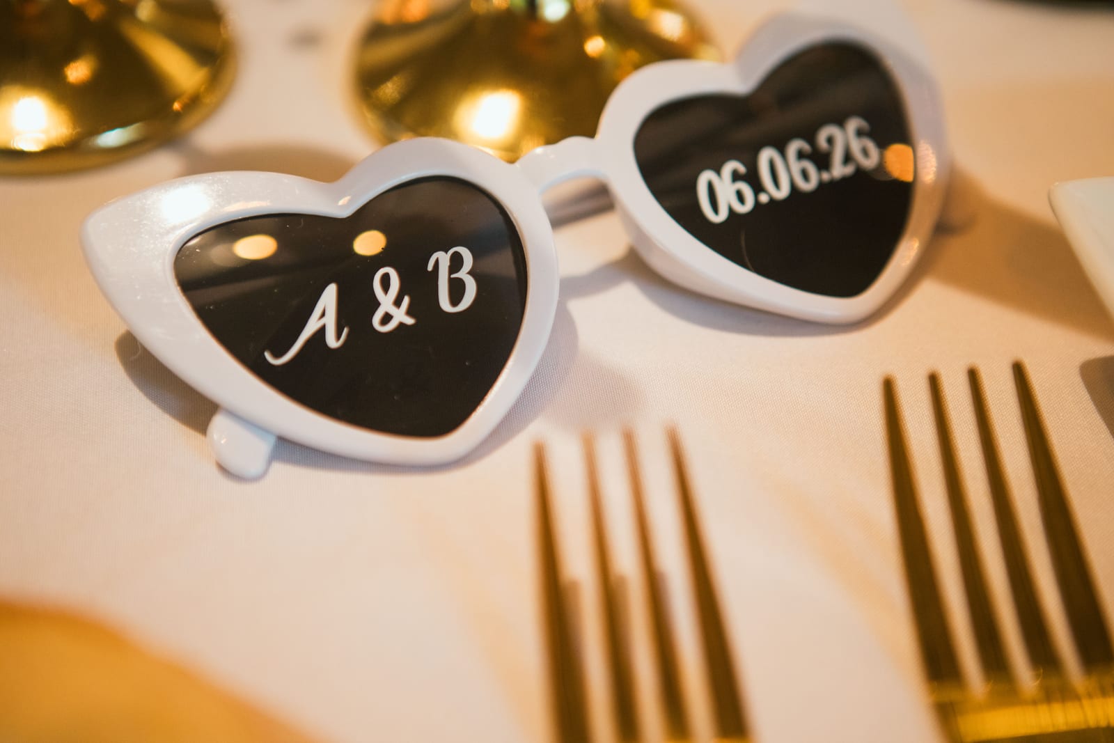 Lunettes de soleil en forme de cœur personnalisées "A & B" et "06.06.26" posées près des couverts dorés, mariage au Domaine de Senercy, par Jean-Baptiste Argyre (Jaysphotography)