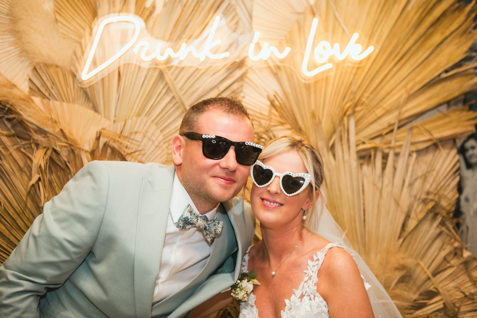 Amandine et Benjamin en lunettes de soleil posent au photo booth devant l'enseigne néon "Drunk in Love" et les palmes séchées, mariage au Domaine de Senercy, par Jean-Baptiste Argyre (Jaysphotography)