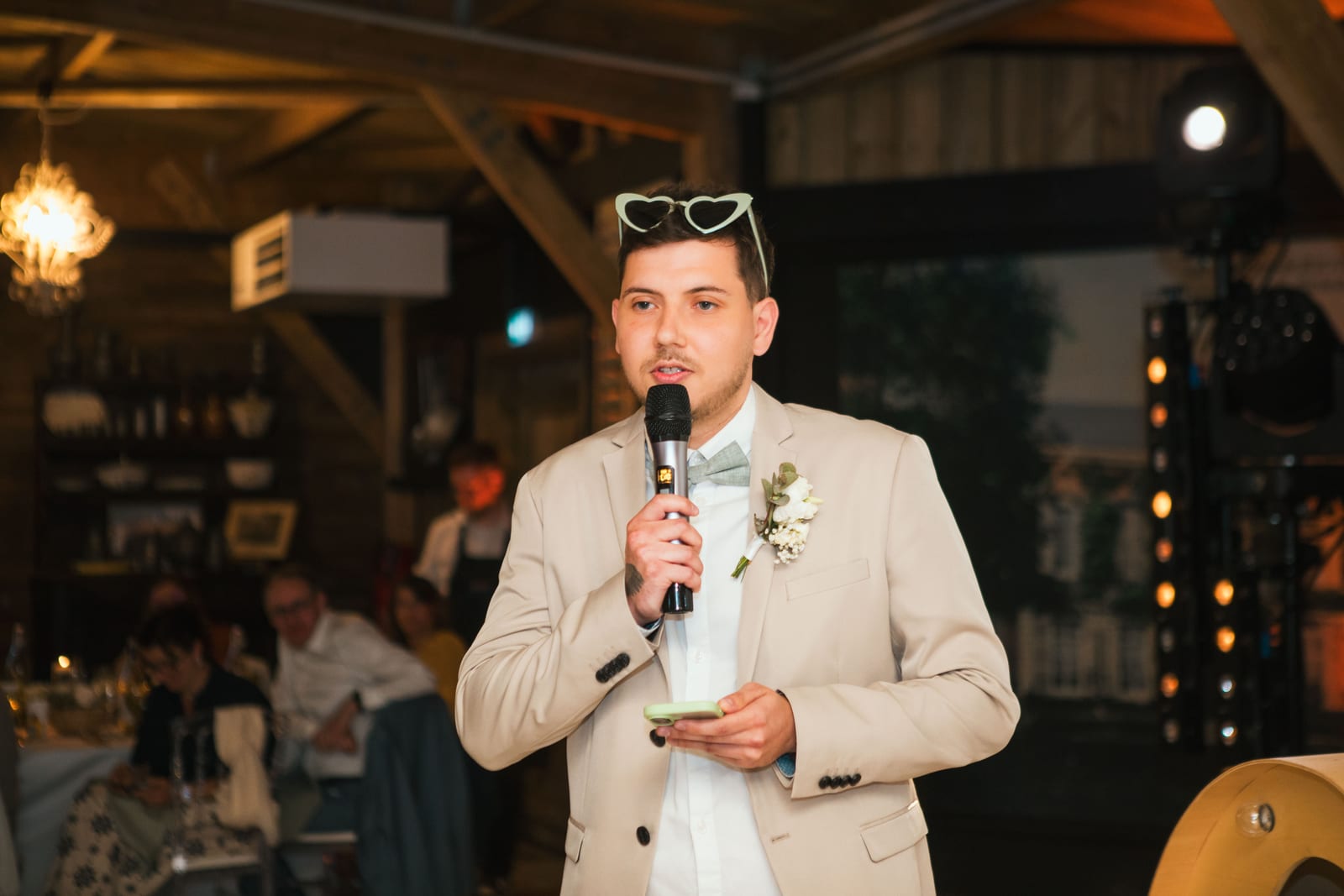 Un témoin en costume beige prononce un discours au micro pendant la soirée, lunettes en cœur sur la tête, mariage au Domaine de Senercy, par Jean-Baptiste Argyre (Jaysphotography)