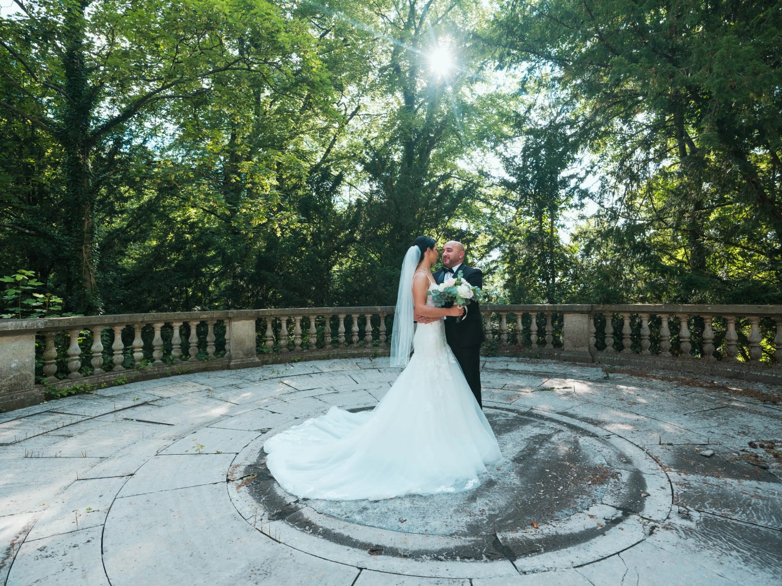 Les mariés enlacés au centre d'une terrasse ronde à balustres sous les grands arbres du parc, soleil entre les feuilles, mariage au Château de Montvillargenne, Gouvieux, par Jean-Baptiste Argyre (Jaysphotography)